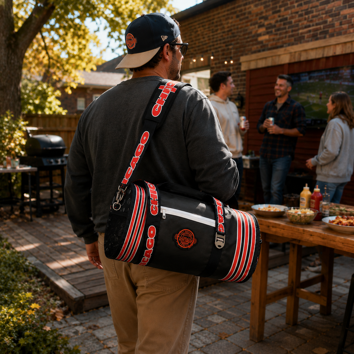 A man wearing a Chicago cap carries a navy Chicago Bulls shoulder duffel bag with red and white stripes and the team logo, in an outdoor backyard setting with people gathered around a grill and tables.